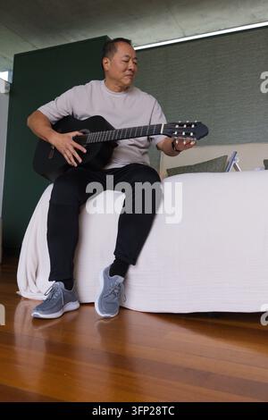 Senior Asian man sitting on bed tuning acoustic guitar in bedroom with tablet wearing gray sneakers Stock Photo