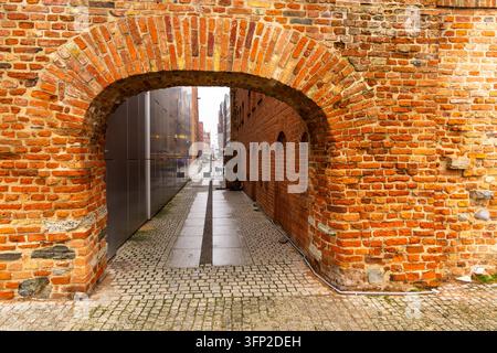 View of a historic red brick archway leading into a narrow cobblestone alleyway with modern architecture in the background in Gdansk, Pomeranian Voivo Stock Photo