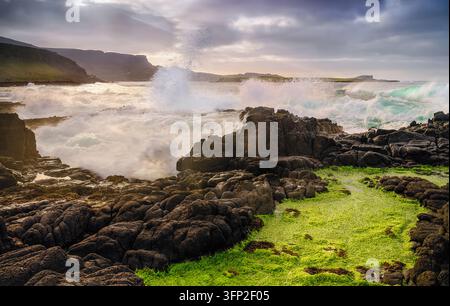 View of the rugged coastline of Skye with crashing waves hitting dark volcanic rocks covered in vibrant green algae under a dramatic sky in Highland C Stock Photo