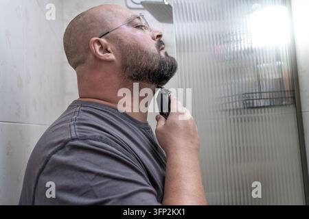 Bearded man performs neckline touchup with electric clipper under chin in bathroom, removing neck stubble between barber visits for tidy appearance and DIY maintenance. Stock Photo