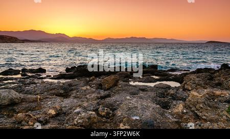 Golden sunset over a rocky beach with waves and distant mountains near Pacheia Ammos in eastern Crete, Greece Stock Photo
