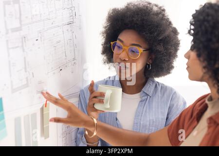 Diverse female designers studying pinned floor plans with color cards in studio, holding mug. Blueprint, architect, collaboration, brainstorming, workspace, modern, bright Stock Photo