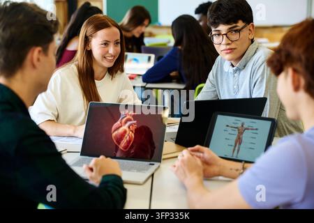 Diverse students engaging in a biology lesson, studying human anatomy on laptops and tablets in a modern classroom Stock Photo
