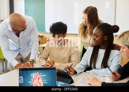 Diverse group of students and male teacher discussing human anatomy, collaborating on a biology project in a modern classroom Stock Photo
