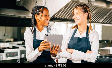 In a big professional kitchen two famous chefs discuss video blog using a tablet. Professionals smiling standing reading searching modern technology a Stock Photo