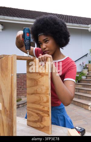 Woman wearing red tee holding plywood while using cordless drill on patio table with tape measure Stock Photo
