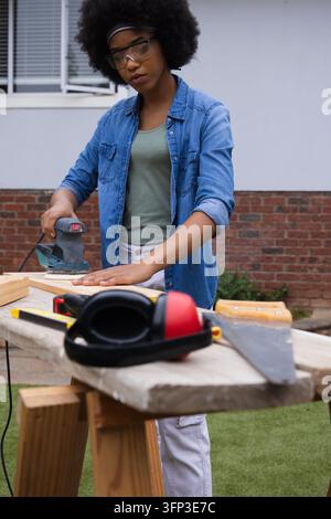 Orbital sander is smoothing wooden plank on portable workbench in backyard near brick wall. Woodworking, diy, outdoor, tools, safety, goggles, bench Stock Photo