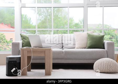 Energy independence and remote work, portable power station charging laptop in a modern home office with large windows and comfortable living room sof Stock Photo