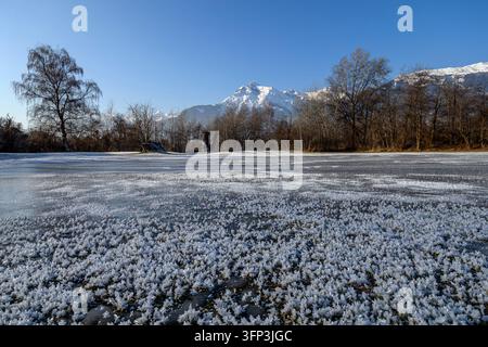 Close-up details of delicate frost crystals on a frozen lake surface with snow-covered alpine mountains Stock Photo