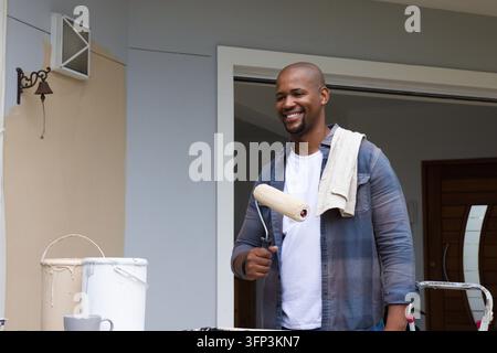 African American man standing on porch holding paint roller, painting wall with bucket, towel Stock Photo