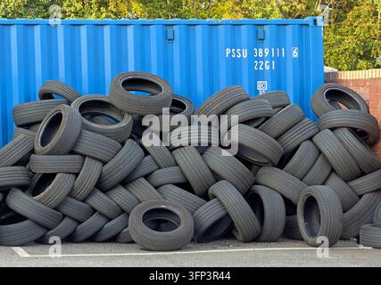 Dozens of discarded black vehicle tyres are stacked irregularly outdoors in front of a blue steel shipping container with trees beyond. Stock Photo