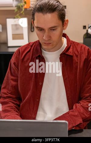Focused mid adult man in burgundy corduroy shirt and white t-shirt works on a laptop at a dark desk in a cozy home office. Concept of remote work, dee Stock Photo