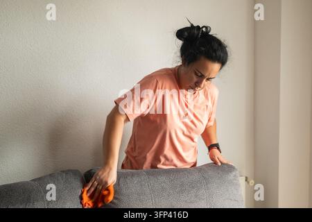 Focused mid adult woman in a peach t-shirt wipes the back of a gray fabric couch with an orange microfiber cloth, a routine housework and home hygiene Stock Photo