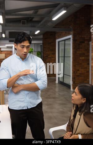 Diverse coworkers with man in blue shirt gesturing and speaking at modern office, white workstation Stock Photo