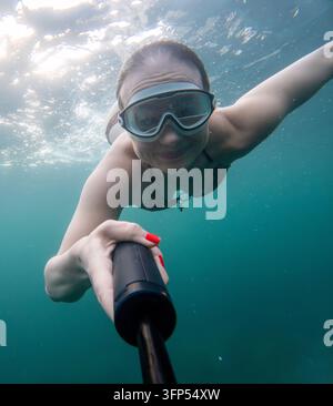 Underwater selfie, smiling young woman swimming with goggles in clear sea holding action camera stick Stock Photo
