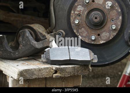 A close-up view shows a set of brand new brake pads positioned on a rustic wooden surface, adjacent to a worn and corroded vehicle brake caliper and r Stock Photo
