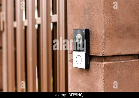 •  Close-up of a modern black electronic video doorbell camera installed on a brown wall near a fence. Smart home security system, wireless entrance s Stock Photo