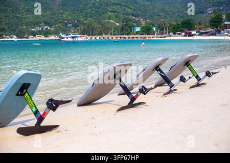 Several efoil surfboards lie lined up on a white sandy beach near clear turquoise ocean water. Modern water sports activity setup on Koh Phangan tropi Stock Photo
