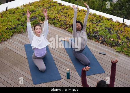 Diverse females in activewear practicing yoga kneeling on rooftop deck with blue mats, water bottle Stock Photo