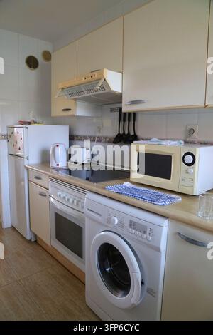 Generic basic normal kitchen interior in Spain. Kitchen with fridge, oven, microwave, washing machine and wall mounted overhead cabinets. Stock Photo