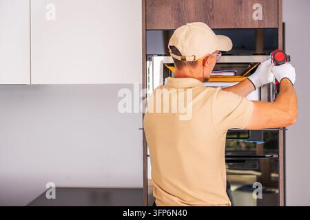 Rear view of a worker using a cordless drill to secure an open built in oven in tall wood grain kitchen cabinetry. Stock Photo
