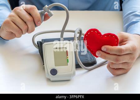 Close view of person using digital blood pressure monitor while holding red heart shape, illustrating cardiovascular health and medical care Stock Photo