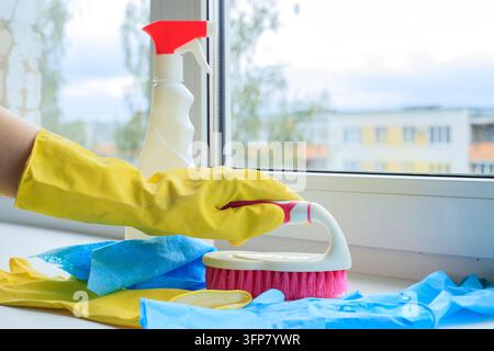 Gloved hand holding scrubbing brush with spray bottle on window sill for domestic cleaning concept. Household hygiene equipment and detergent supply o Stock Photo