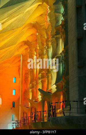 Warm orange and cool green stained glass light washes over sculpted stone columns and the vaulted ceiling above a curved walkway inside the Sagrada Fa Stock Photo