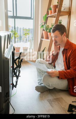 Man sits on the floor with a tv and wall mount, reading a manual with a confused and frustrated expression while attempting a complex home improvement installation Stock Photo