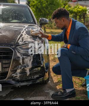 Young man in blue suit carefully scrubs foamy black car hood outdoors, showing hands on vehicle cleaning, shine restoration, and polished maintenance Stock Photo