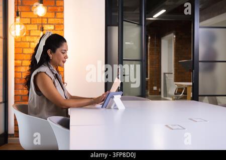 Woman in sleeveless vest sitting left of conference table holding paper and using tablet Stock Photo
