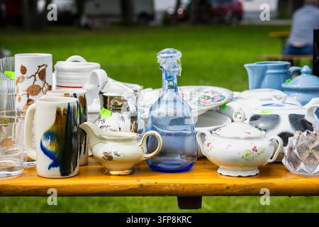 Vintage tableware displayed at an outdoor flea market, featuring porcelain teapots, ceramic mugs, glassware, a blue glass bottle, floral dishes. Stock Photo