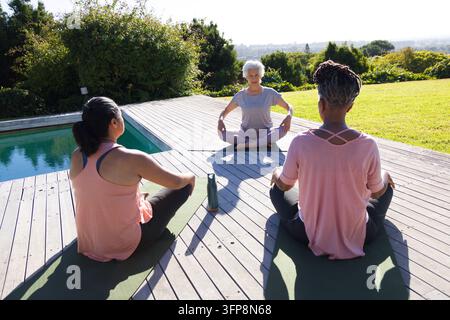 Senior woman leading two women practicing yoga on deck by pool on yoga mats in activewear Stock Photo