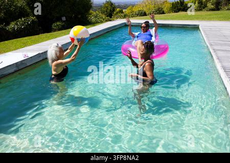 Diverse female friends playing in backyard pool, tossing striped beach ball, lounging on pink ring Stock Photo