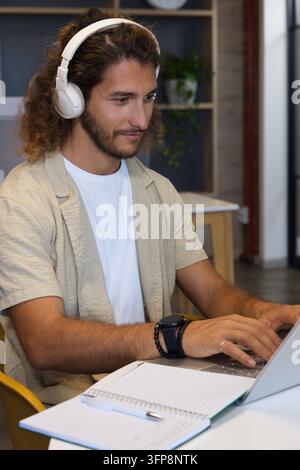 Man typing on laptop while wearing white over-ear headphones at co-working table with notebook Stock Photo