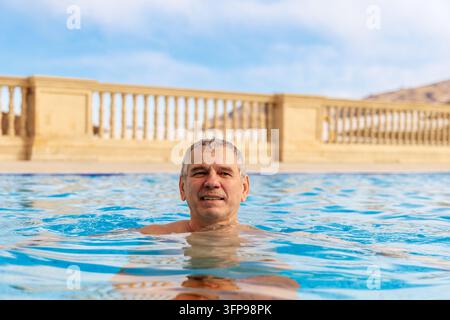 Medium close up of a handsome senior man in a heated outdoor swimming pool at a winter wellness spa hotel looking at camera. Stock Photo