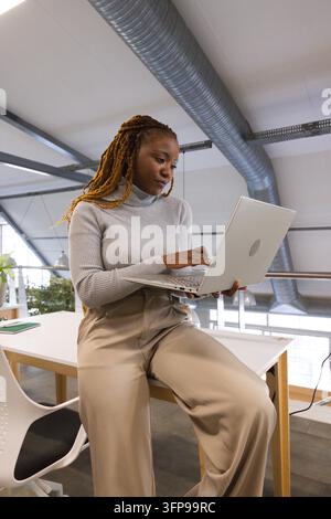 African woman sitting on table edge using silver laptop at modern open-plan office with notebook Stock Photo