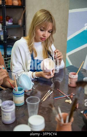Young woman applying glaze to handmade ceramic mug with brush in pottery workshop. Pottery glazing process represents creativity and artisan Stock Photo