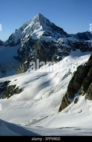 A stunning view showcases the towering peak of Mont Blanc, partially shrouded in sunlight. The surrounding landscape is dominated by vast snowfields a Stock Photo