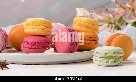 An assorted selection of delicate French macarons in pastel shades of pink, yellow, orange, and green rests on a white ceramic plate, styled with soft Stock Photo