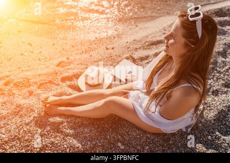 Woman beach laptop young woman sits on pebbly beach using laptop computer during golden hour sunset on summer vacation Stock Photo