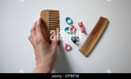 A hand is holding a wooden comb while two wooden combs and several colorful hair ties are placed on a white surface. This scene shows personal groomin Stock Photo