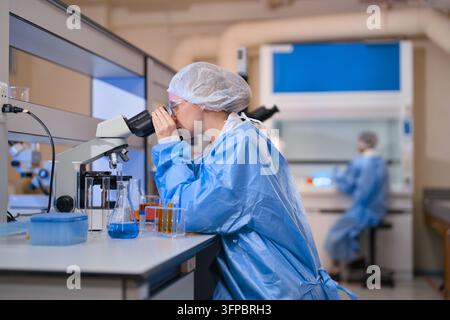 Woman scientist examining samples under a microscope in a clean research lab, surrounded by beakers and equipment, conducting medical and pharmaceutic Stock Photo