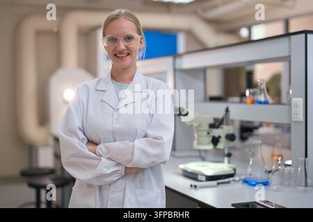 Young female scientist in a white lab coat and safety goggles standing proudly with arms crossed in a modern laboratory, researching chemistry and bio Stock Photo
