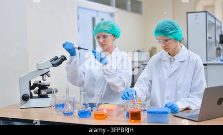 Two female scientists in lab coats and protective eyewear collaborate at a modern bench, using pipettes, microscopes and colorful solutions for chemis Stock Photo