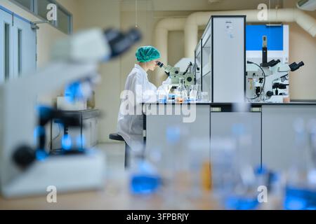 Scientist in a lab coat and hairnet conducting research with a microscope, observing samples with blue and orange liquid chemicals in glassware, symbo Stock Photo