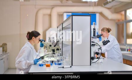 Two female scientists in a laboratory conducting scientific research, examining samples under microscopes, collecting data on a digital tablet, and wo Stock Photo