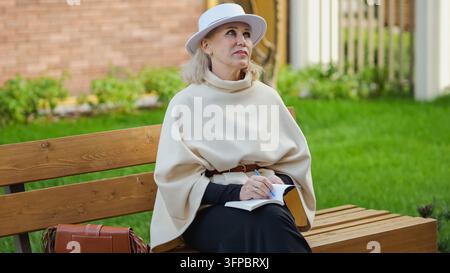 Mature woman writer writes by hand in notebook while sitting outdoors. Reflective author holds blue pen and gazes upward, finding inspiration beside Stock Photo