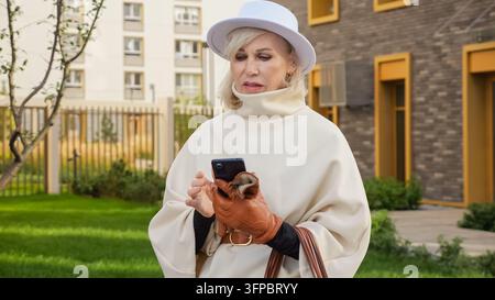 Elegant older woman checks smartphone messages outdoors, wearing white hat and coat with brown gloves. Woman studies her mobile with focused, slightly Stock Photo
