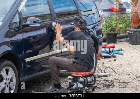 A detailing technician wearing a headlamp sits on a rolling stool while meticulously polishing the exterior door of a blue vehicle with a rotary buffi Stock Photo